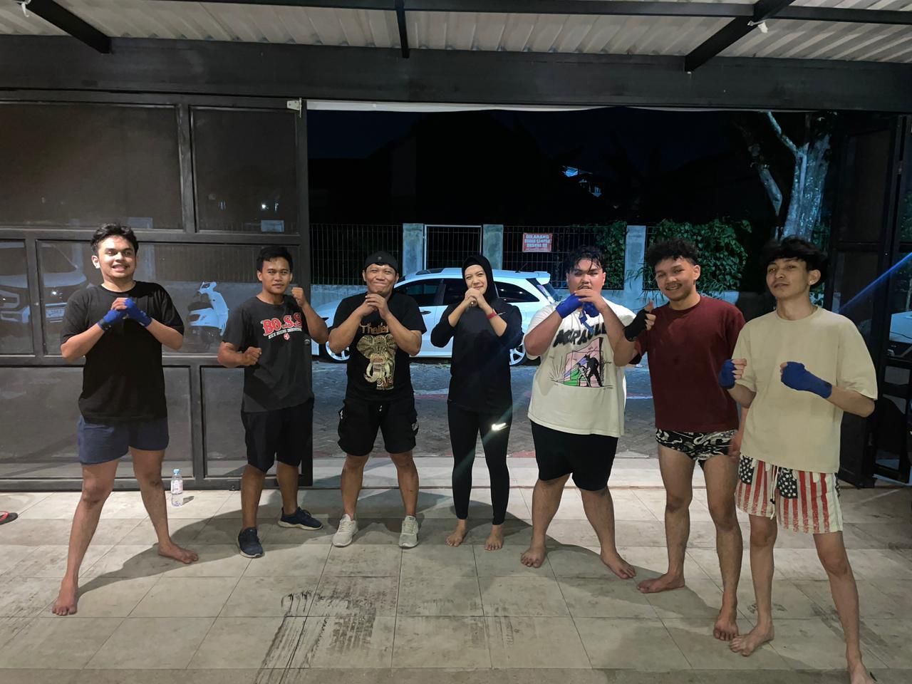 Tujuh penghuni berpose kuda-kuda muay thai dengan sarung tangan di area depan kos pada malam hari